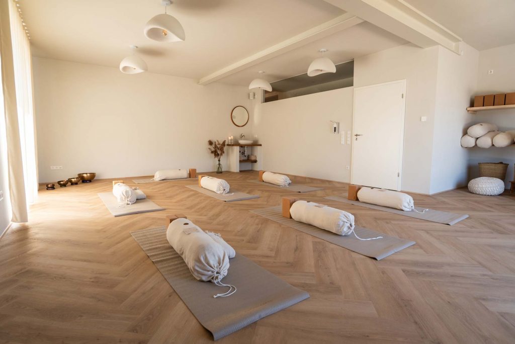 Lichtdurchfluteter, gemütlicher Yogaraum mit ruhiger Atmosphäre – Blick auf die ausgelegten Yogamatten und vorbereiteten Kissen, ready für die Yoga-Stunde.