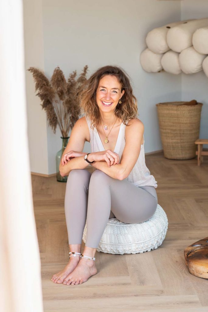 Christin Rupp sitzt lächelnd auf einem Meditationskissen im Yogabereich – entspannte Sitzhaltung, freundliche Begrüßung